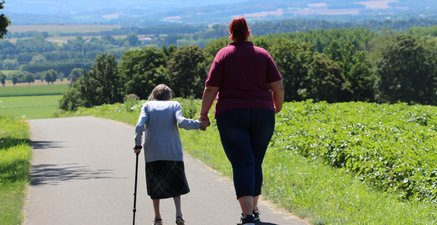 Das Bild zeigt eine kleine Oma an der Hand einer großen Betreuungskraft. Sie gehen spazieren.
