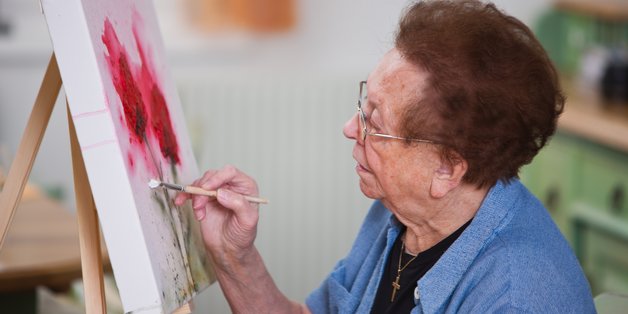 Eine alte Frau steht vor einer Staffelei und malt konzentriert.