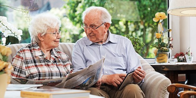Alter Mann und alte Frau sitzen auf dem Sofa. Sie liest Zeitung, er schaut in ein Tablet.