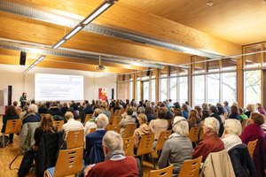 Teilnehmende der BAGSO-Fachtagung folgen einem Vortrag in einem hellen Konferenzraum. Links im Vordergrund steht eine Referentin am Rednerpult, rechts steht ein Banner der BAGSO.