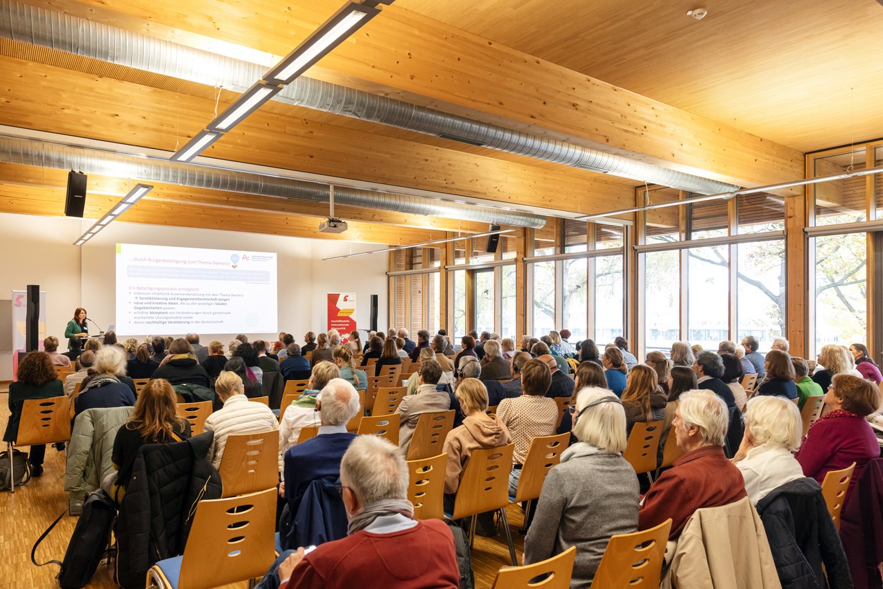 Teilnehmende der BAGSO-Fachtagung folgen einem Vortrag in einem hellen Konferenzraum. Links im Vordergrund steht eine Referentin am Rednerpult, rechts steht ein Banner der BAGSO.