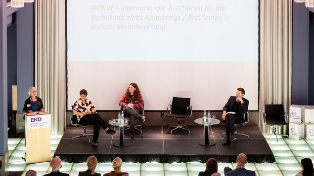 Links, an einem Rednerpult hält eine Frau eine Ansprach. Rechts davon stehen vier Sessel für eine Diskussionsrunde. Dort sitzen bereits ein Mann und zwei Frauen. Klick öffnet eine vergrößerte Ansicht.