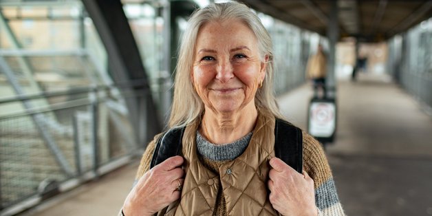 Ältere Frau mit langen grauen Haaren. Sie trägt einen Rucksack auf dem Rücken. Sie steht und im Hintergrund sind digitale Anzeigetafeln zu sehen.