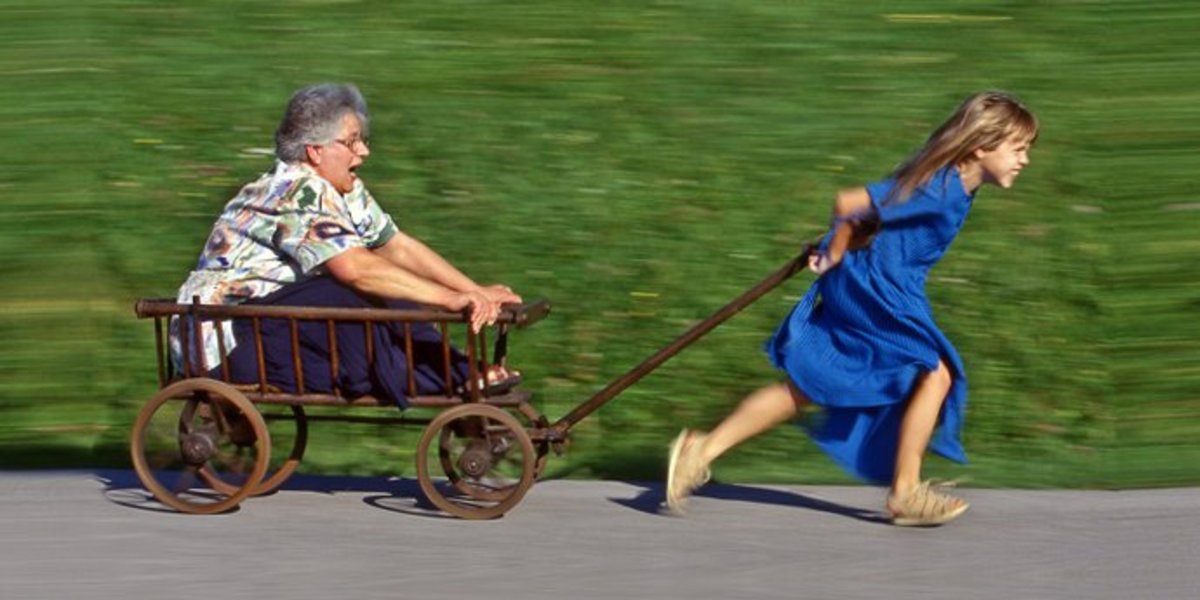 Mädchen im blauen Sommerkleid läuft und zieht dabei einen Leiterwagen, in dem eine ältere Frau sitzt