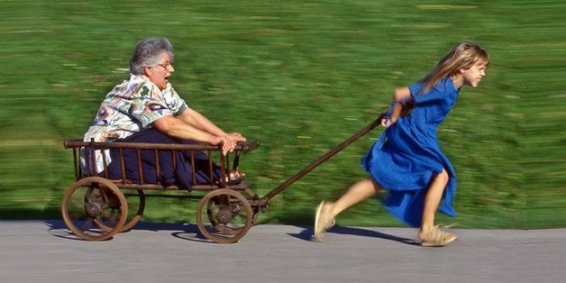 Mädchen im blauen Sommerkleid läuft und zieht dabei einen Leiterwagen, in dem eine ältere Frau sitzt
