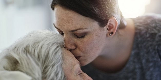 Ältere Dame im Bett. Eine jüngere Frau beugt sich zu ihr und gibt ihr einen Kuss auf die Stirn 