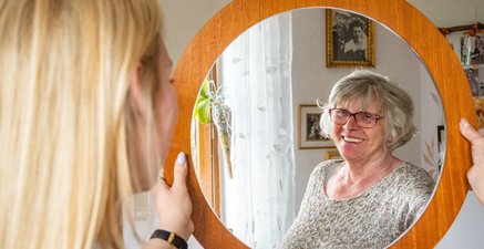 Unscharfer Hinterkopf einer jungen Frau, in der Mitte ein runder Spiegel, das Spiegelbild einer älteren Frau