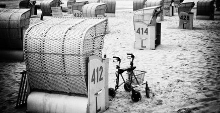 Ein Rollator steht neben einem Strandkorb am Meer. Es ist eine Schwarzweiß-Aufnahme.