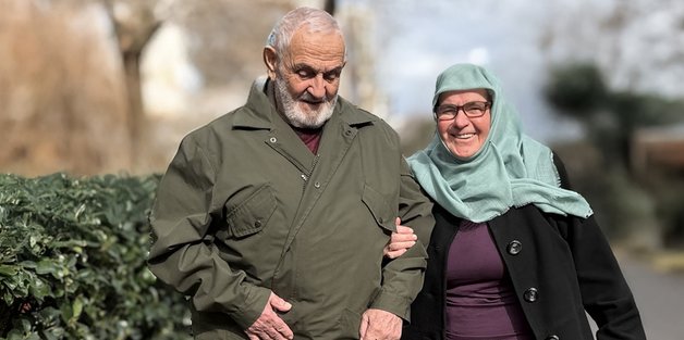 Ein alter Mann und eine alte Frau gehen auf einem Bürgersteig, entlang einer Lorbeerhecke. Sie trägt einen dunklen Mantel, hat eine helles Kopftuch an und hat sich bei dem Mann untergehakt. Er trägt einen grünen Parka. Er hält mit der einen Hand die Jacke zu.