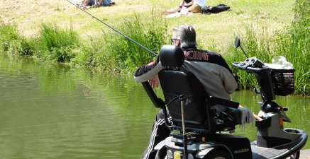 Mann sitzt auf E-Rollstuhl angelnd an einem Teich. Auf der anderen Uferseite sitzen junge Frauen auf einer Wiese.