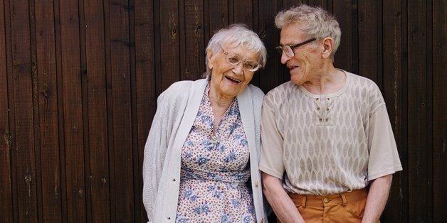 Ältere Frau und älterer Mann stehen an einer Holzwand.