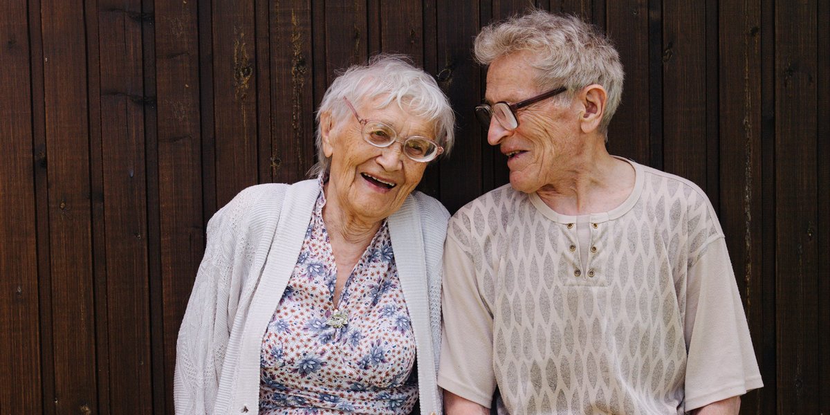 Ältere Frau und älterer Mann stehen an einer Holzwand.