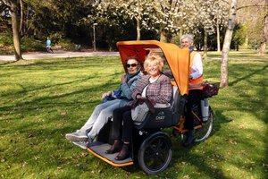 In einem Park steht eine Riksha. Zwei ältere Frauen sitzen darin und zeigen Freude. Hinter ihnen sitzt die Radlerin. Auch eine ältere Frau mit kurzen grauen Haaren. Sie trägt eine orangefarbene Weste.
