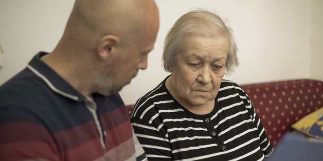 Ein Mann mit Glatze sitzt neben einer alten Frau auf einem Sofa. Er zeigt ihr etwas auf einem Klemmbrett, das er in der Hand hält.