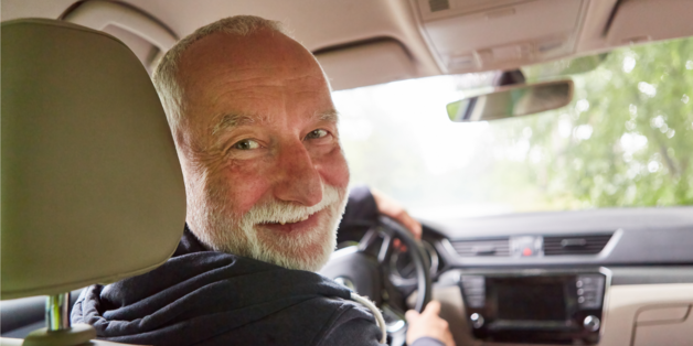 Ein älterer Autofahrer im Inneren eines Auto. Er schaut lächelnd nach hinten. 