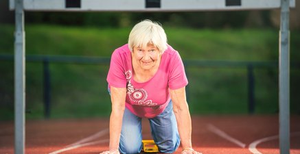 Eine alte Dame sitzt im Startblock vor zwei Hürden auf einer Laufbahn, den Blick nach vorne in die Kamera.
