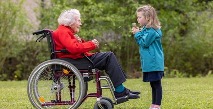 Seniorin im Rollstuhl, ihr gegenüber ein kleines Mädchen, das ihr eine Pusteblume zupustet