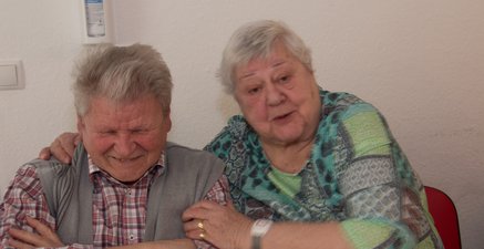 Frau I re. hält Herrn H seitlich im Arm. Sie lacht. Er verschränkt die Arme und schaut lachend nach unten