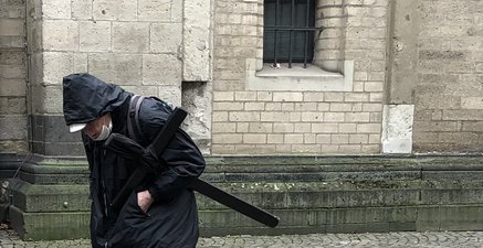 Alter schlanker Mann, dunkel gekleidet, Kapuze ins Gesicht gezogen, trägt schwarzes Holzkreuz unterm Arm