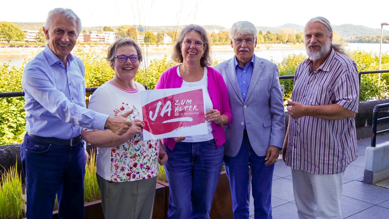 Zwei Frauen und drei Männer stehten draußen. Im Hintergrund sieht man eine Fluss. Eine Frau hält ein Schild in der Hand mit der Aufschrift "Ja zum Alter!" Klick öffnet eine vergrößerte Ansicht.