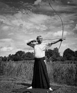 Älterer Mann mit Brille und japanischer alter Tracht steht in der freien Natur. Er hält einen gespannten Bogen in der Hand. Es ist Kyudo, eine japanische Kunst des Bogenschießens.