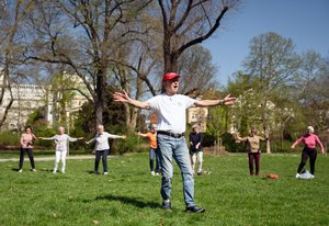 Ein älterer Mann in verwaschener Jeans, mit weißem T-Shirt, Turnschuhen und einer roten Kappe auf dem Kopf hat die Arme weit ausgebreitet und ruft etwas. Er steht in einem Park. Hinter ihm stehen Frauen, die Gymnastik machen.