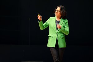 Eine Frau mittleren Alters, Ernährungsdoc Silja Schäfer, steht auf der Bühne. Sie hat schulterlanges schwarzes Haar, trägt eine Brille und einen sehr auffällig grünen Blazer. Sie hält eine kleine blaue Flasche in der Hand und schauf darauf.