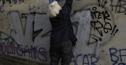 Ältere Frau steht auf Zehenspitzen vor einer Wand und übersprüht eine Hassbotschaft