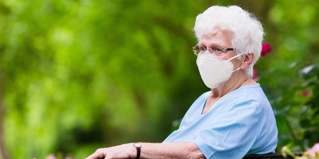 Eine alte Frau mit einer FFP2-Maske sitzt im Garten auf einer Bank