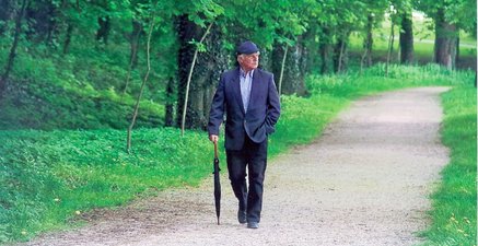 Älterer Mann mit Kappe und Regenschirm geht auf einem Waldweg spazieren