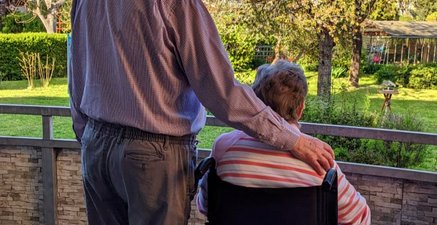 Ein altes Ehepaar auf dem Balkon. Sie sitzt im Rollstuhl. Er steht neben ihr und legt seinen Arm auf ihre Schulter.