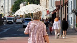 Alte Frau läuft bei Sonnenschein durch eine Stadt. Sie hat einen Schirm aufgespannt.