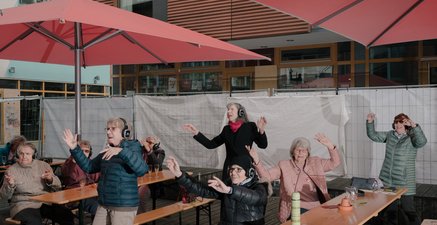 Sechs ältere Frauen tragen Kopfhörer und tanzen. Während ein paar sitzen, stehen andere. Sie bewegen ihre Hände.