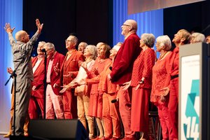 Ein Chor auf der Bühne: Alte Frauen und Männer, in Rottönen gekleidet singen gemeinsam. Vor ihnen steht der Chorleiter. Ein Mann mittleren Alters mit Glatze. Er is komplett in grau gekleidet. Er schaut den Chor an und hat seine Arme nach oben gestreckt.