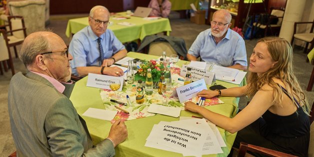 Im Veranstaltungsraum sind mehrere Tische mit neongrünen Tischdecken, an denen Menschen sitzen, die diskutieren. Am Tisch im Vordergrund sitzen drei alte Männer und eine junge Frau, die miteinander diskutieren.