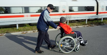 Älterer Mann schiebt seine Frau im Rollstuhl, wärend im Hintergrund ein ICE vorbei fährt.