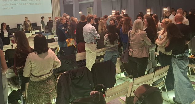 Blick in den Saal. Die meisten Personen stehen beieinander und unterhalten sich. Klick öffnet eine vergrößerte Ansicht.