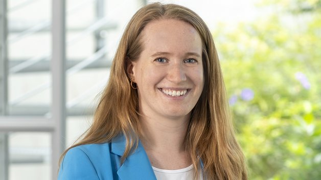 Protrait Marie Großmann: Junge Frau mit langen braunen Haaren. Sie trägt ein weißes T-Shirt und darüber einen hellblauen Blazer. Sie lächelt mit strahlenden Augen.