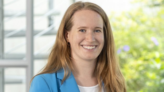 Protrait Marie Großmann: Junge Frau mit langen braunen Haaren. Sie trägt ein weißes T-Shirt und darüber einen hellblauen Blazer. Sie lächelt mit strahlenden Augen.