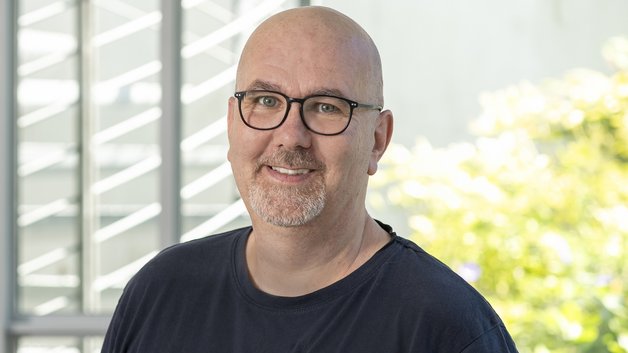 Portrait Klemens Lenz: Mann mittleren Alters mit Glatze, Brillenträger. Er trägt ein schwarzes T-Shirt. Er schaut lächlelnd.