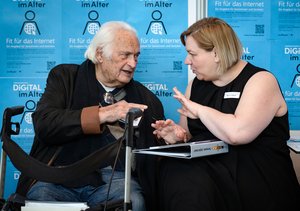 Ein alter Mann mit schwarzer Jacke und Jeans. Vor ihm steht ein Rollator. Eine junge Frau neben ihm, hat einen Aktenordner auf ihrem Schoß. Darauf steht "Startpaket Digitall dabei". Auch sie ist schwarz gekleidet. Sie schauen sich in die Augen und sind in einem intensiven Gespräch.