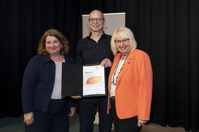 Zwei Frauen, ein Mann. Der Mann steht in der Mitte und hält eine Urkunde in der Hand. Klick öffnet eine vergrößerte Ansicht.