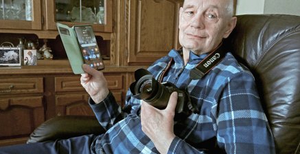 Älterer Mann sitzt in einem Ledersessel, Smartphone in der rechten Hand, Fotokamera um seinen Hals.