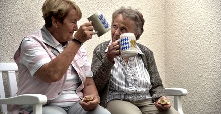 Eine ältere und eine hochaltrige Frau sitzen auf einer Bank und prosten einander mit Tonkrügen zu.