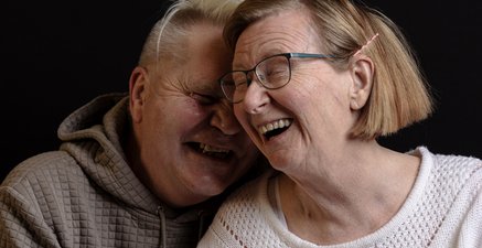 Portrait von zwei Menschen vor schwarzem Hintergrund, beide lachend. Einer ist Pfleger, die andere Seniorin