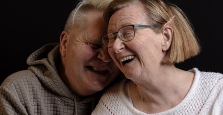 Stärker Zusammen Portrait von zwei Menschen vor schwarzem Hintergrund, beide lachend. Einer ist Pfleger, die andere Seniorin
