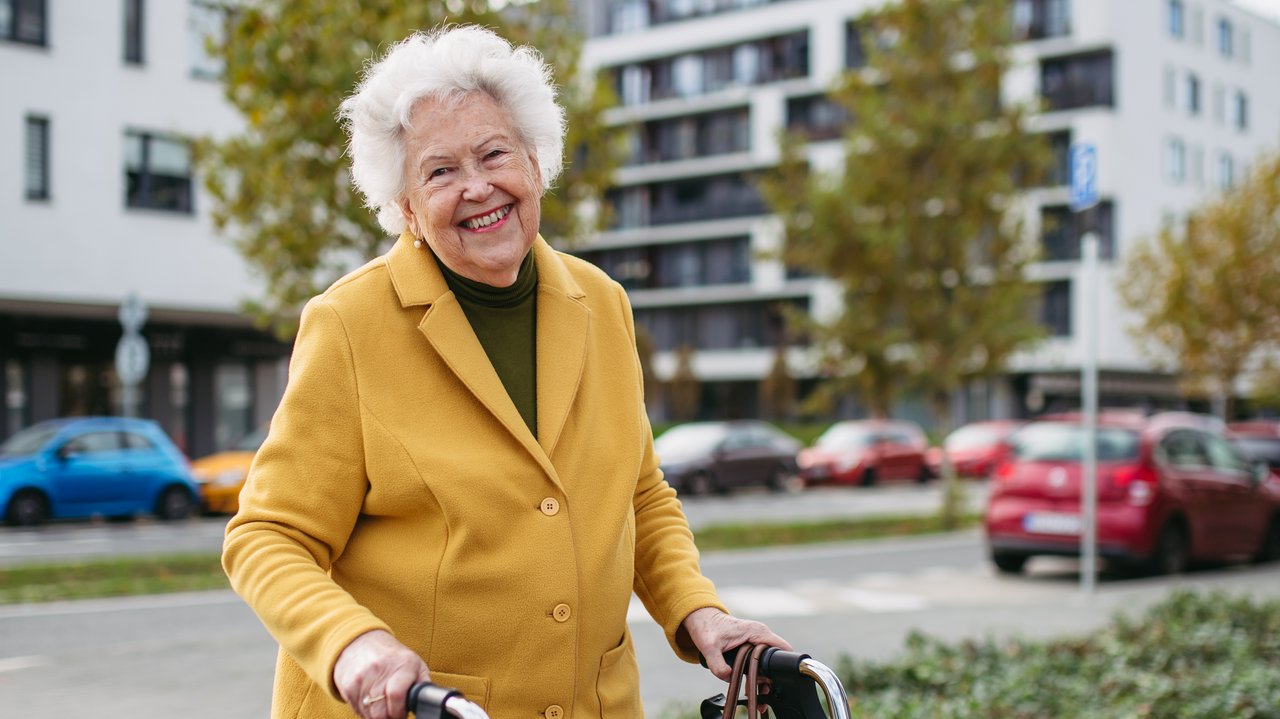 Eine alte Frau läuft mit Ihrem Rolator über den Bürgersteig und lächelt. Im Hintergrund sieht man Häuser und eine Straße, auf der Autos fahren.