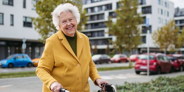 Eine alte Frau läuft mit Ihrem Rolator über den Bürgersteig und lächelt. Im Hintergrund sieht man Häuser und eine Straße, auf der Autos fahren.