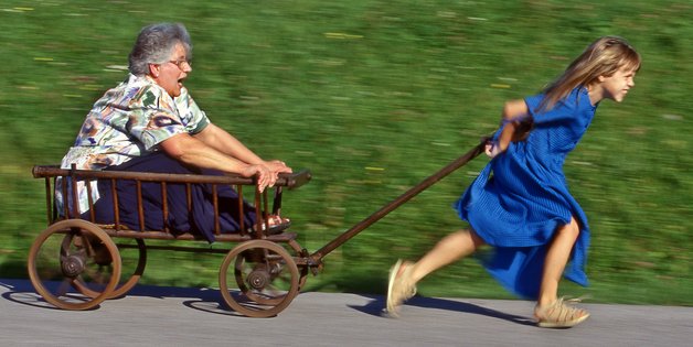 Mädchen zieht einen Bollenwagen, in dem eine ältere Frau sitzt. Beide haben großen Spaß.