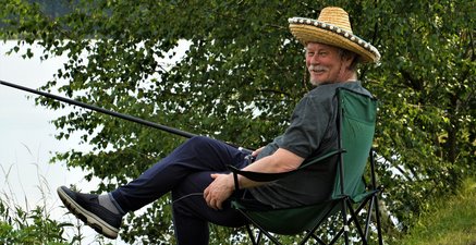 Ein älterer Mann sitzt an einem See mit einer Angel in der Hand. Er trägt einen Strohhut und lächelt in die Kamera.